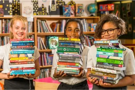 Group of three children each holding a pile of books in a school library setting.